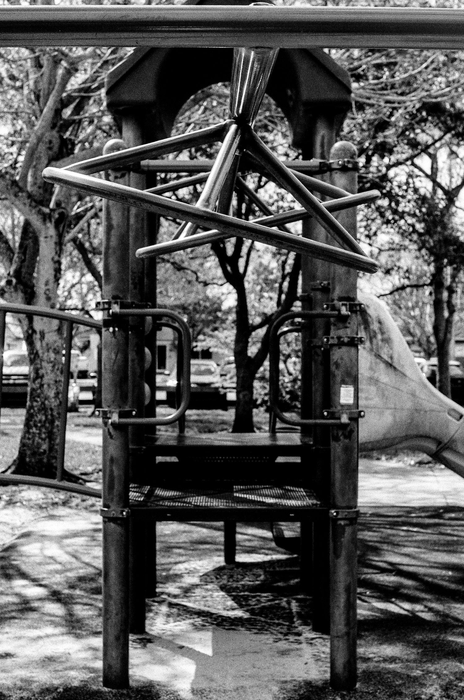 A black and white photograph of vintage playground equipment with metal bars and climbing structures in a park setting.