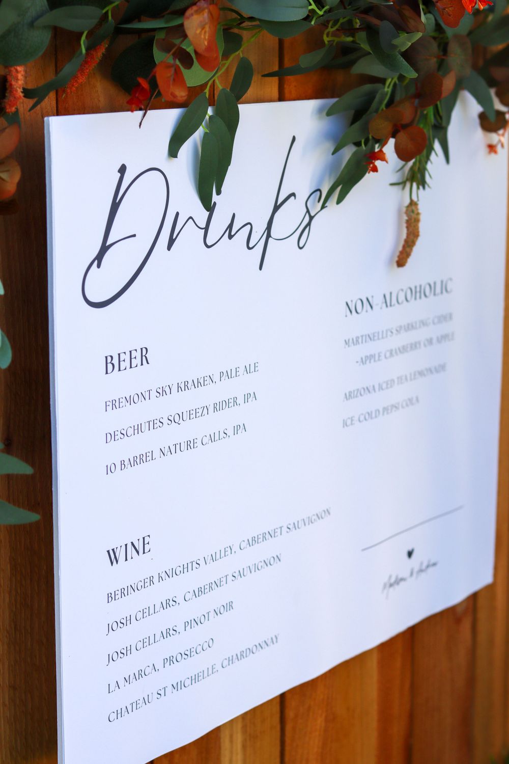 Wedding drinks menu with beer and wine listings hanging against wooden backdrop with foliage.
