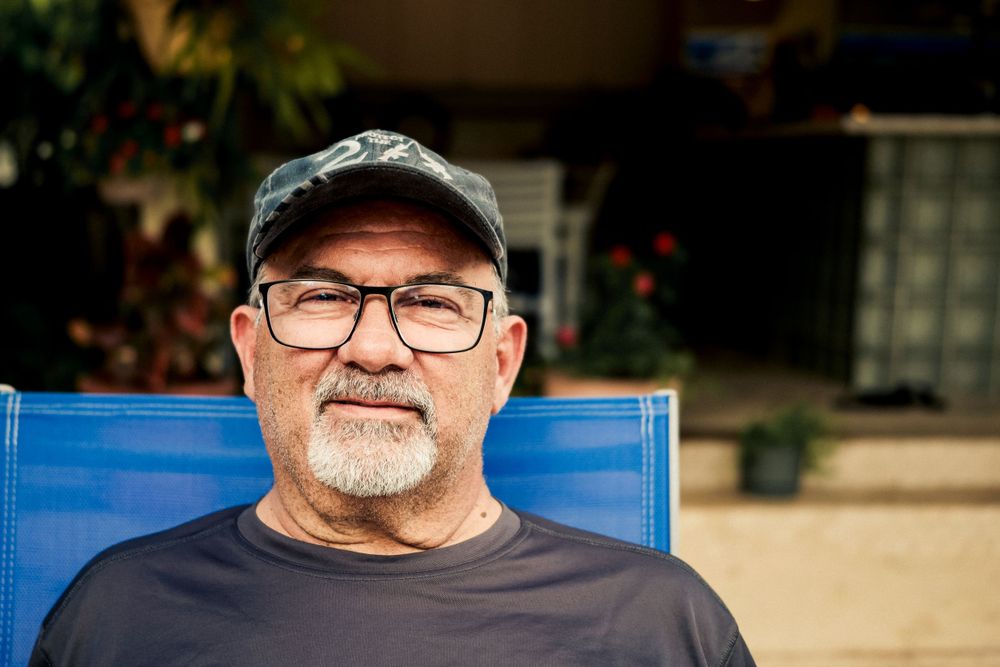 Someone wearing glasses and a baseball cap sits in a blue lawn chair while smiling at the camera.