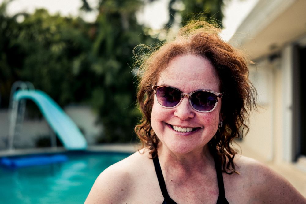 Someone smiling by a swimming pool with sunglasses on a sunny day, with a blue water slide visible in the background.