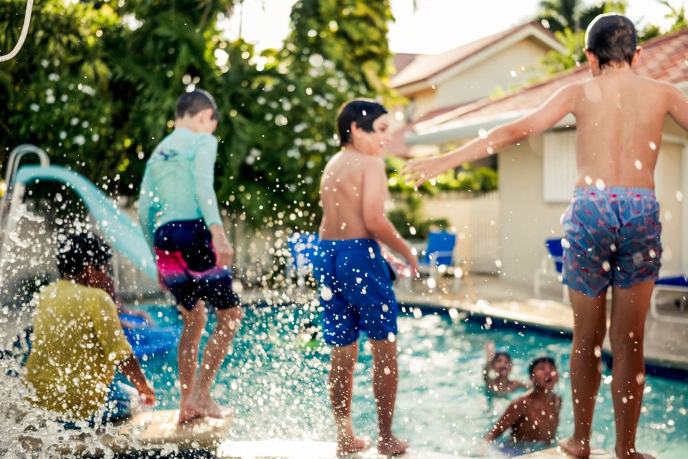 Kids jumping and splashing in a backyard swimming pool on a sunny summer day creating water spray effects.