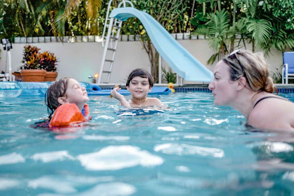 Several people enjoy swimming in a backyard pool with a bright blue water slide on a sunny day.