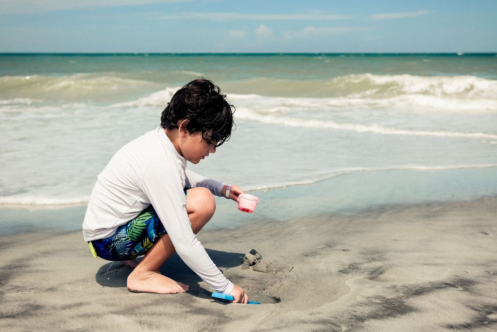 Child wearing a white shirt and blue shorts plays in the sand at the beach with ocean waves in the background.