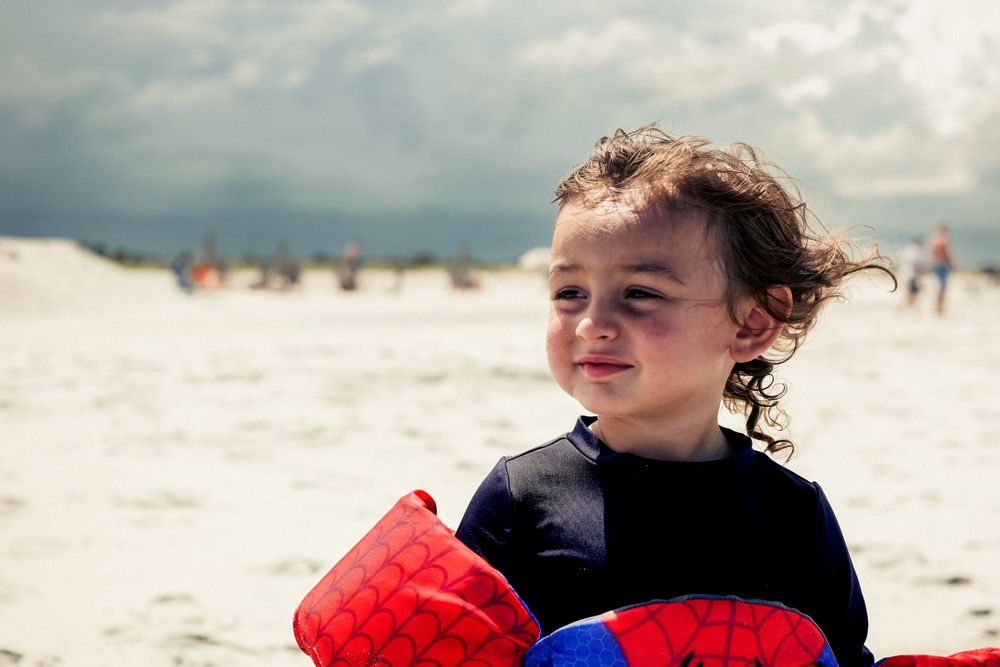 A child in a navy shirt holds a red and blue item while enjoying a sunny day at the white sandy beach.