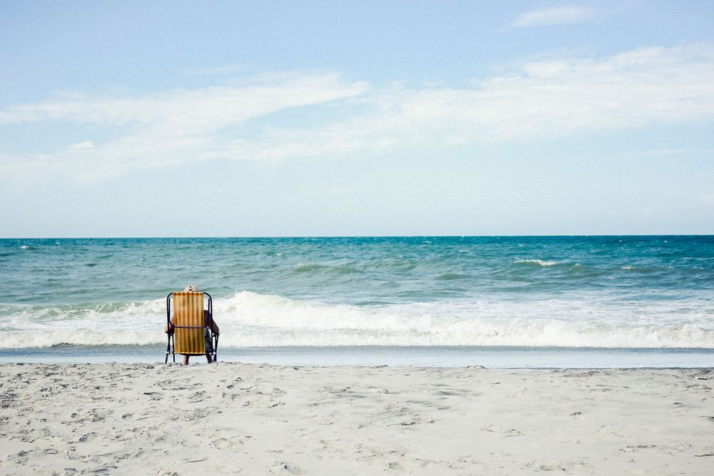 A lone brown wooden beach chair sits empty on a white sandy beach facing the turquoise ocean waves.
