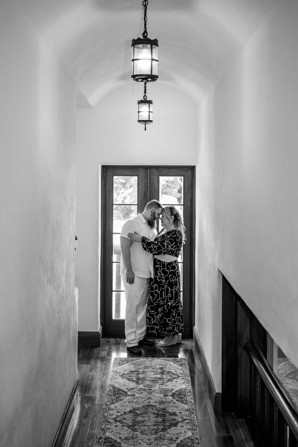 A romantic black and white photograph of two people embracing in a hallway with vintage lanterns overhead.