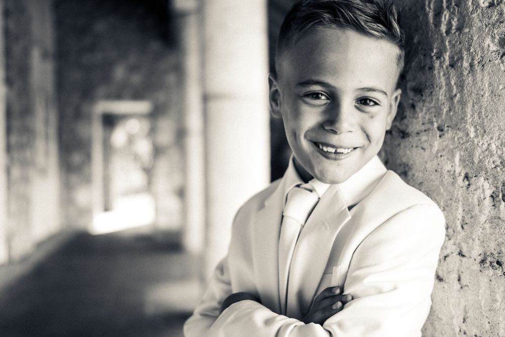 Black and white portrait of young boy in white shirt leaning against wall with arms crossed and smiling expression.
