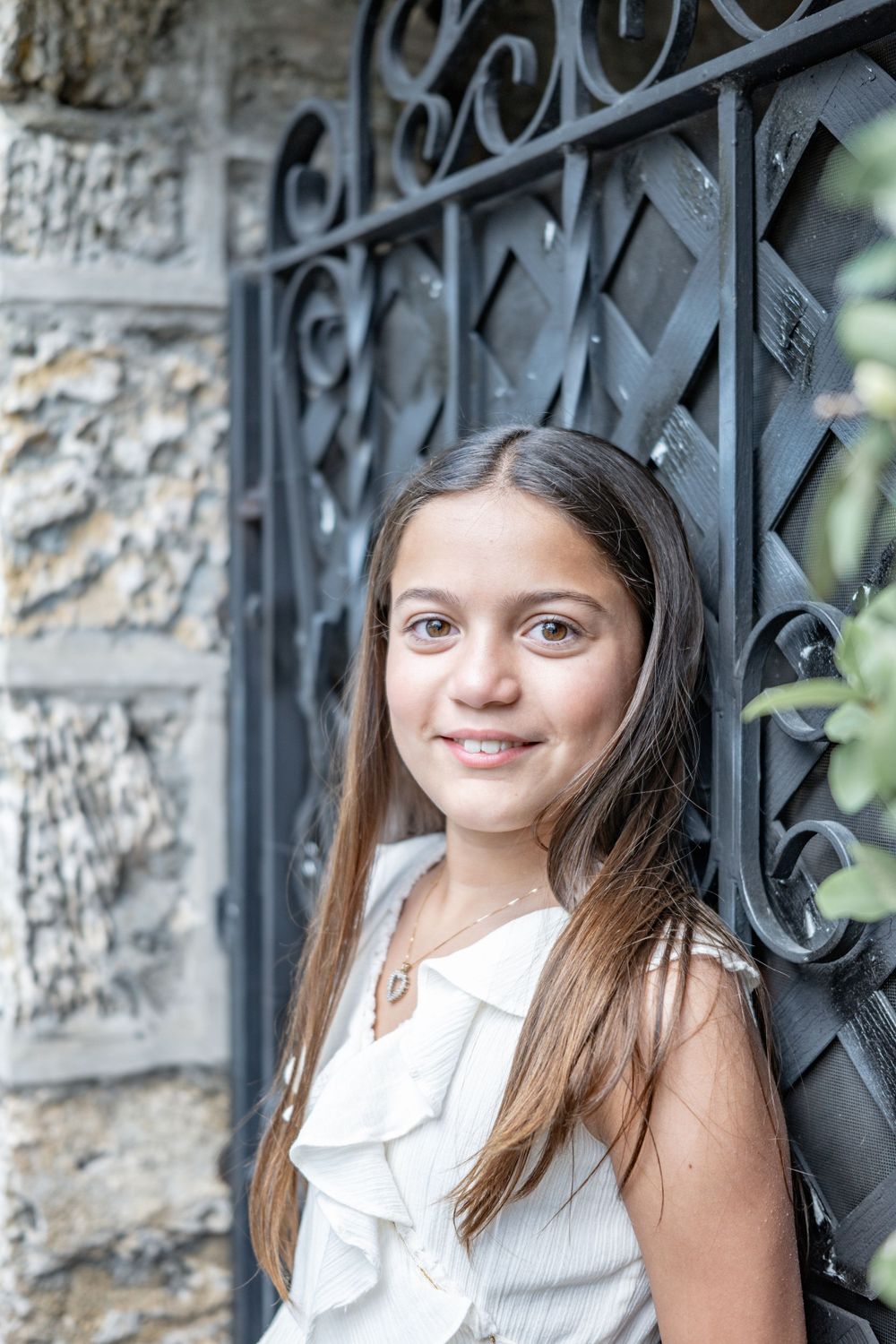 A young girl in a white ruffled top poses against an ornate black iron gate and stone wall background.