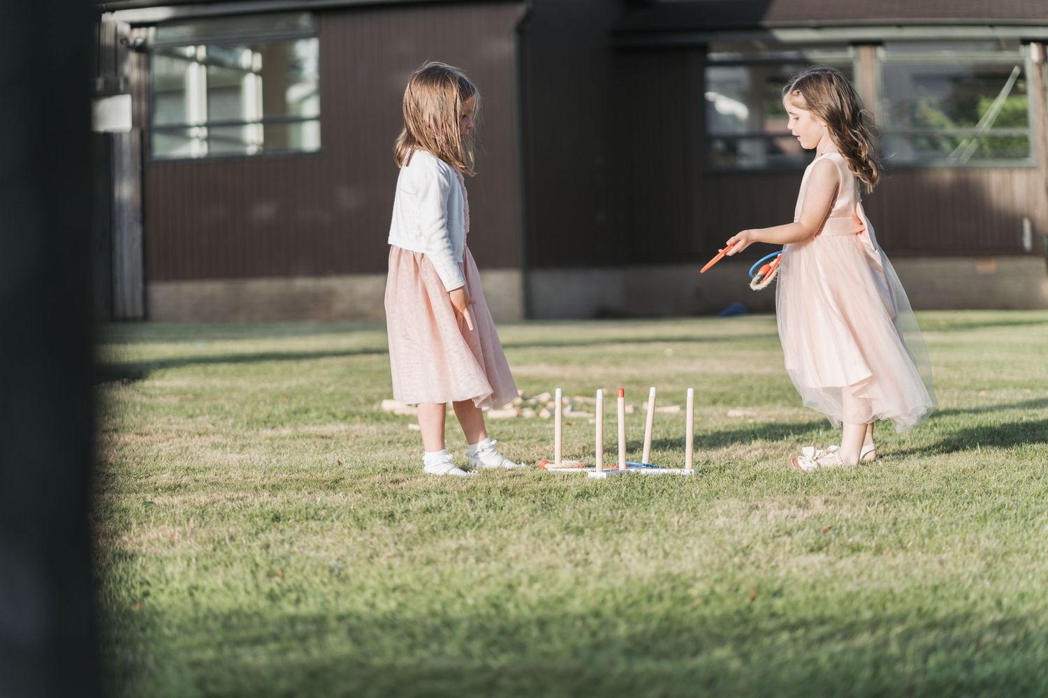 Two children in pink dresses play on a grassy lawn next to a white fence in soft natural lighting.