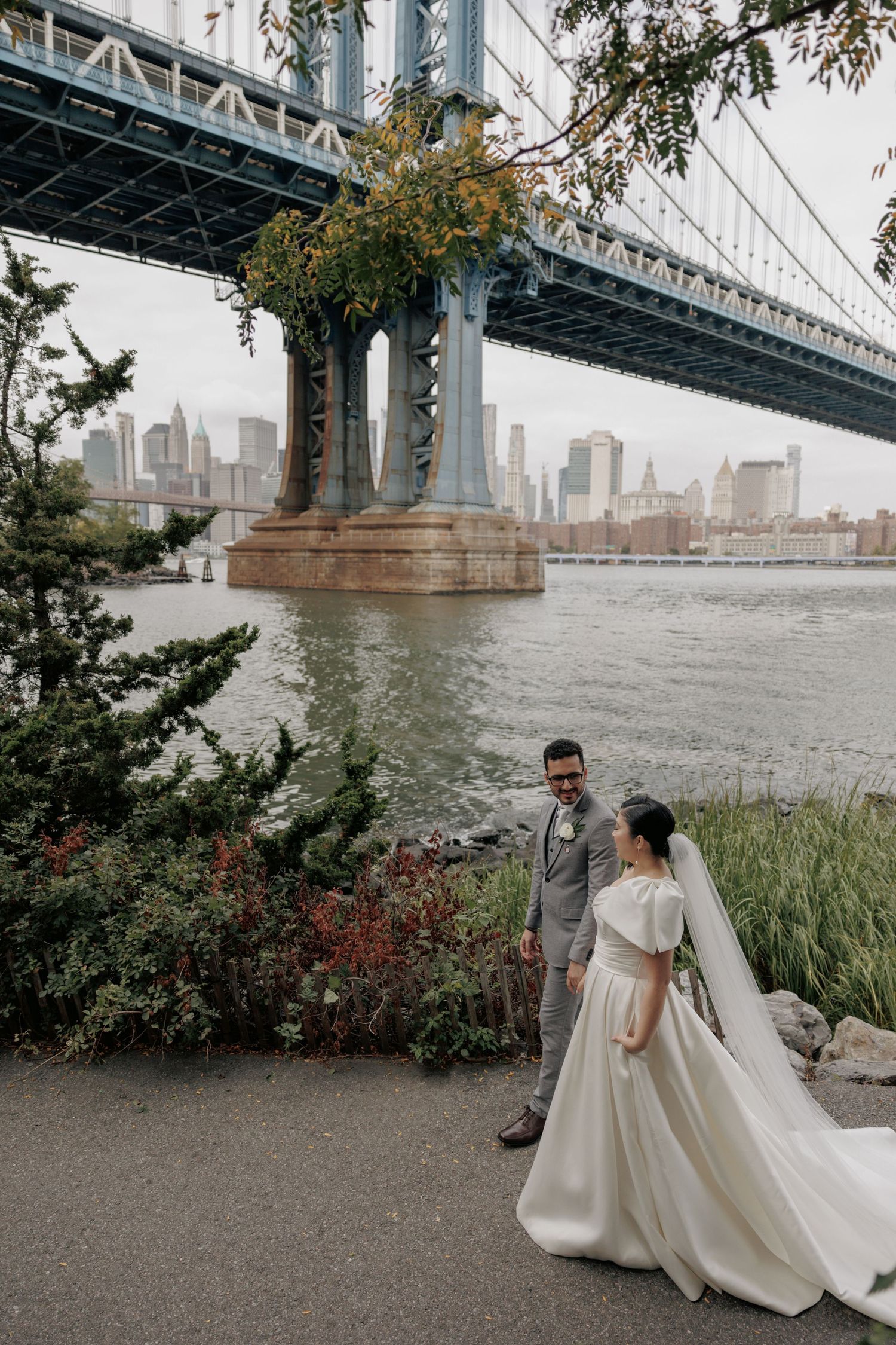 Wedding couple poses beneath Manhattan Bridge with NYC skyline in background.