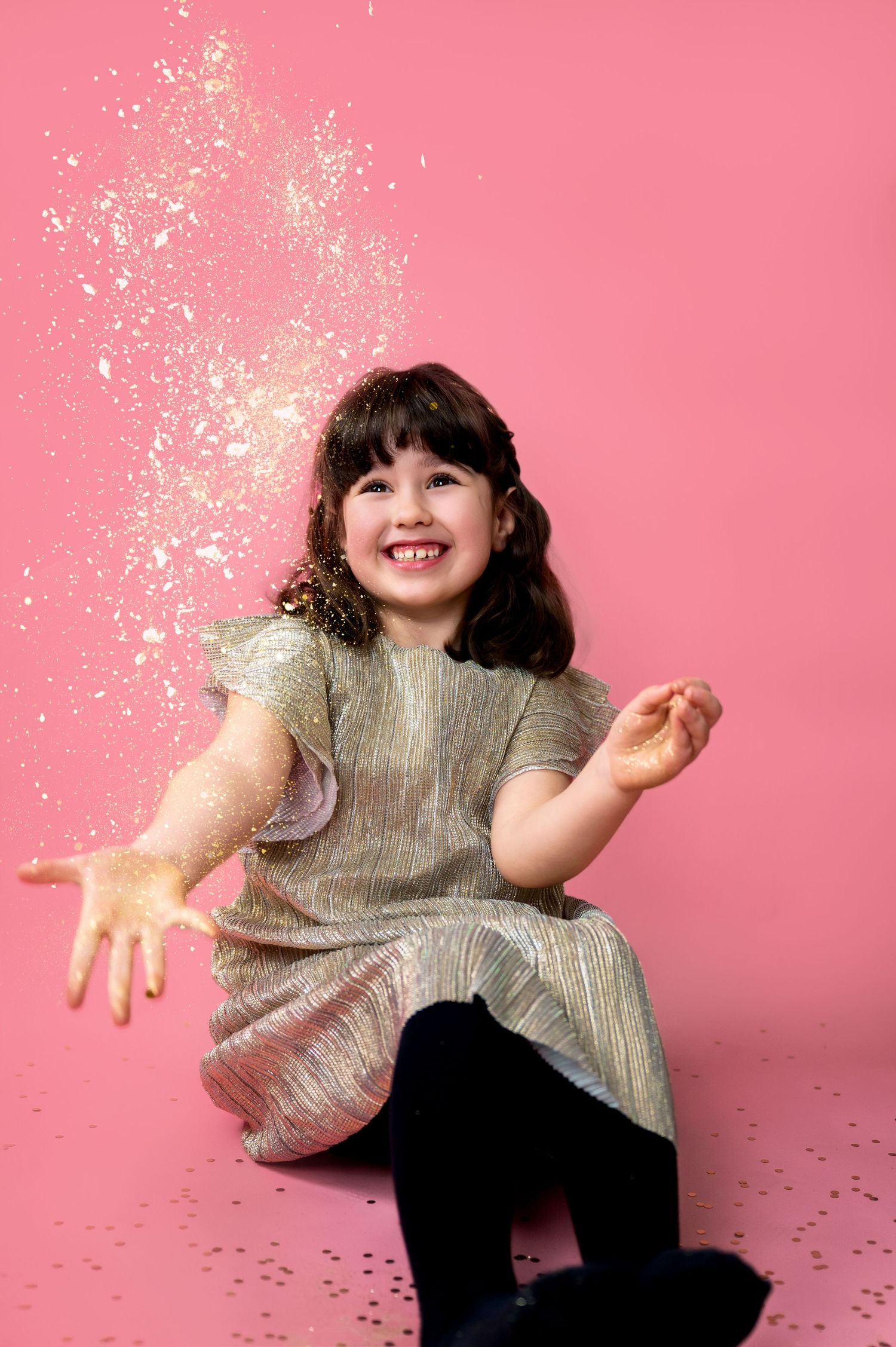 A young child in a metallic dress throws glitter in the air against a pink backdrop while sitting and smiling.