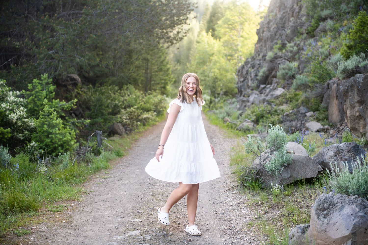 A white sundress twirls on a scenic mountain path surrounded by rocks and greenery in soft evening light.