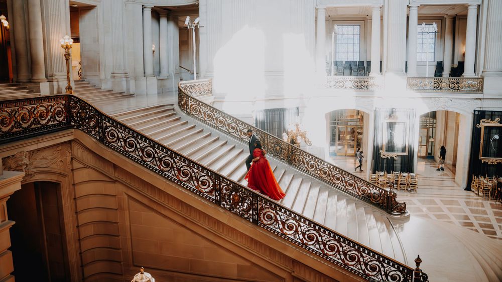 A figure in a flowing red dress stands elegantly on a grand marble staircase in an ornate classical building interior.