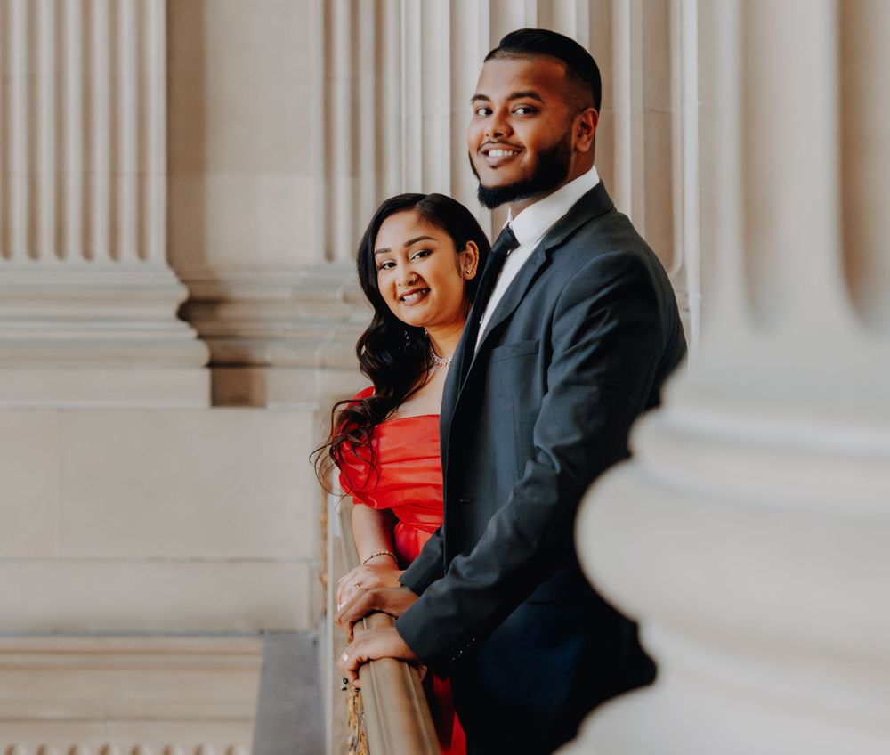 A couple poses together on an elegant staircase, with the woman wearing a red dress and man in a dark suit.