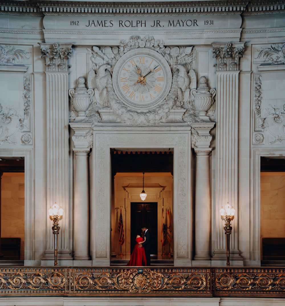 A figure in red stands beneath an ornate clock and architectural details in a grand classical interior.