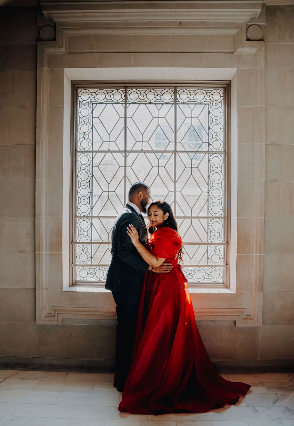 A romantic couple pose against a decorative window, the red dress contrasting with the geometric patterns.