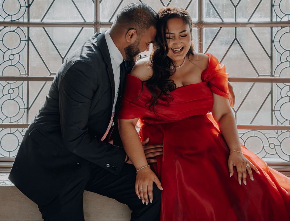 A joyful couple shares a laugh while posing by a patterned window in formal attire.