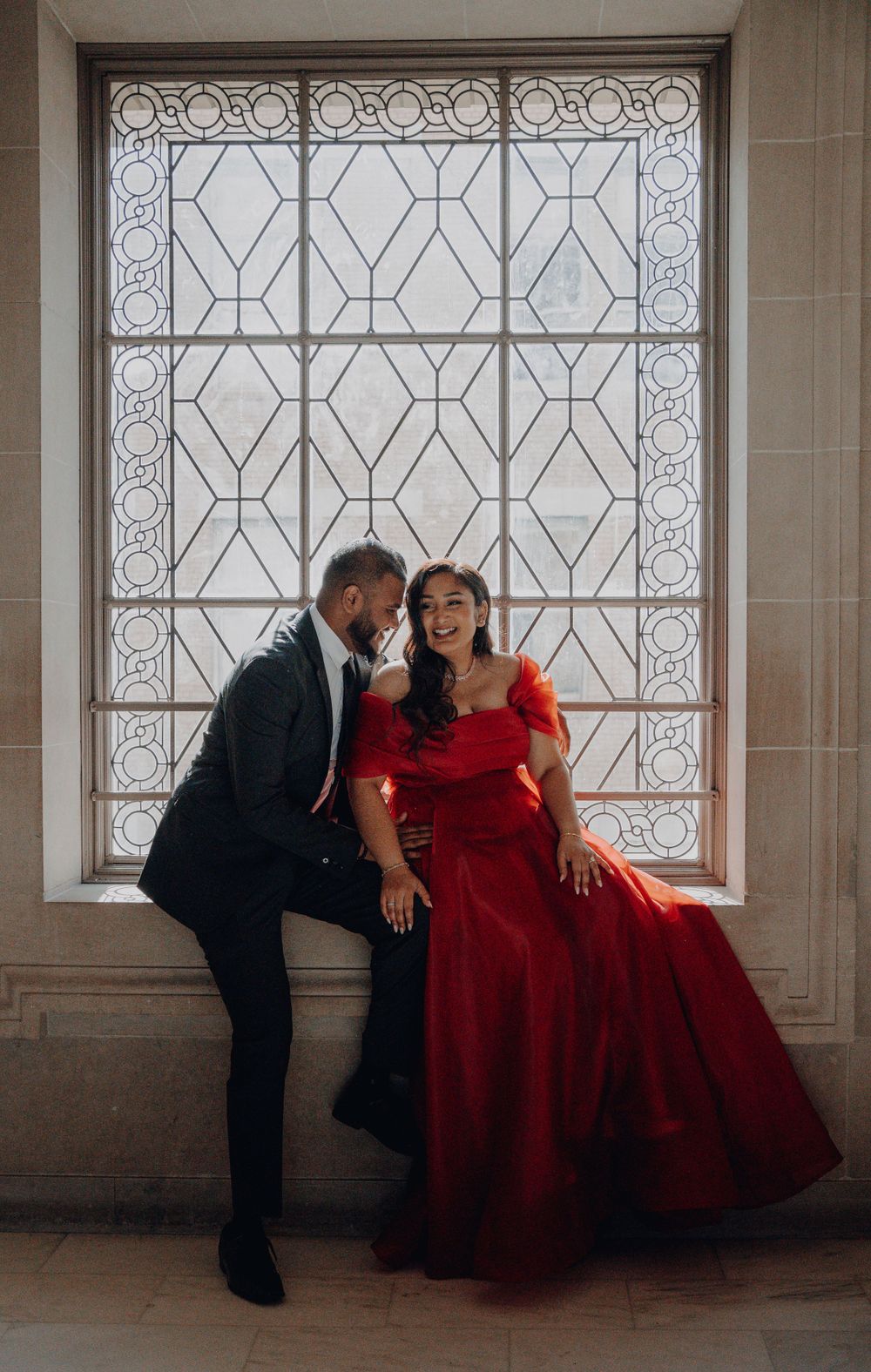 A romantic pose by a decorative window features a stunning red gown and classic black suit.