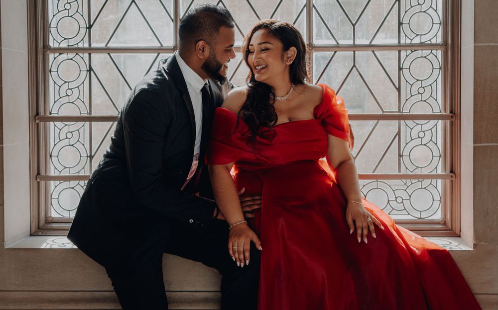 A couple shares an intimate moment by a geometric window, her red dress adding dramatic flair.