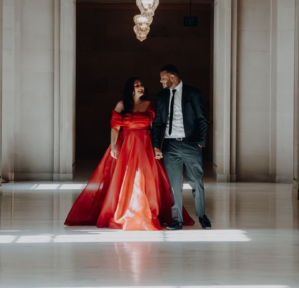 Elegant couple posing in formal attire with a stunning red ball gown and grey suit in a grand hallway.