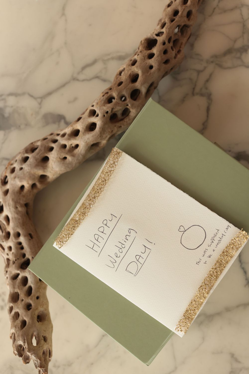Happy Wedding Day card with gold trim rests on a green envelope beside driftwood on a marble surface.
