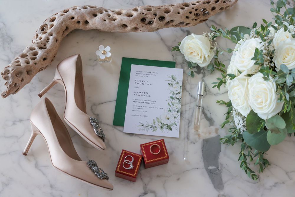 Wedding flat lay featuring nude heels, white rose bouquet, green invitation suite, and ring boxes on marble.
