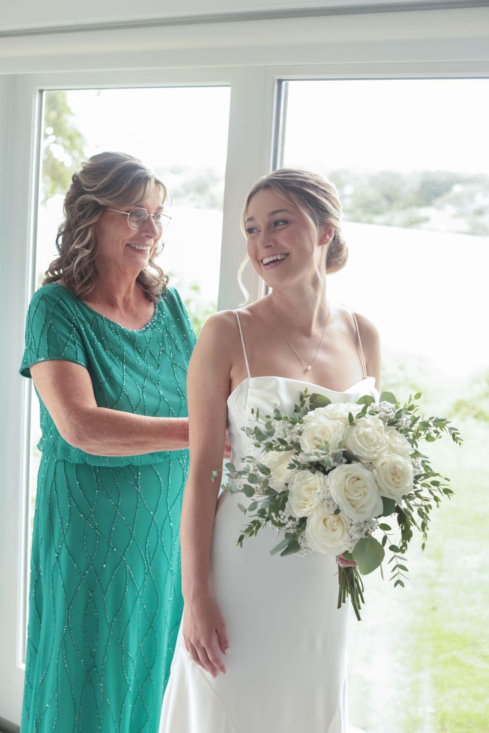 Smiling bride in a white gown holds a white rose bouquet beside her mother in a teal dress by a window.