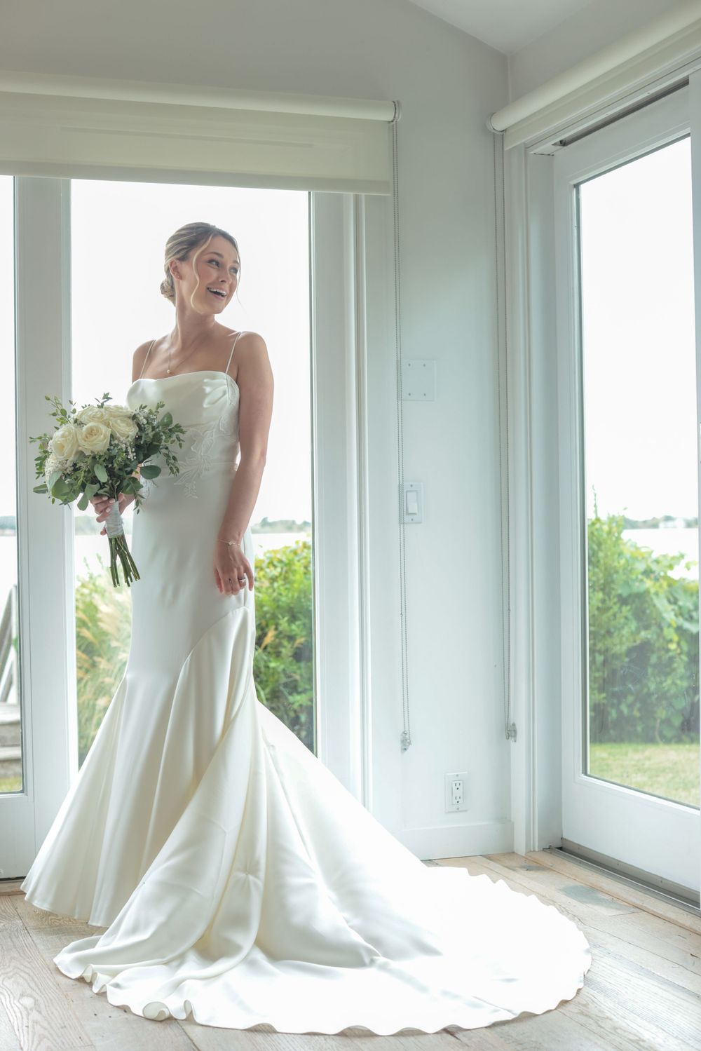 Radiant bride in a fitted white mermaid gown holds a white bouquet while standing by bright windows.