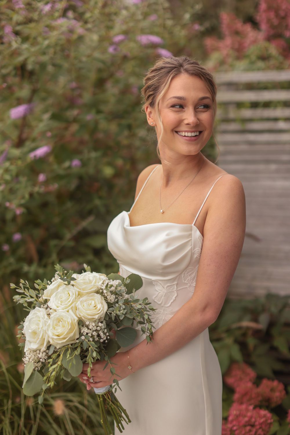 Smiling bride in white satin gown holding white rose bouquet in a garden setting.