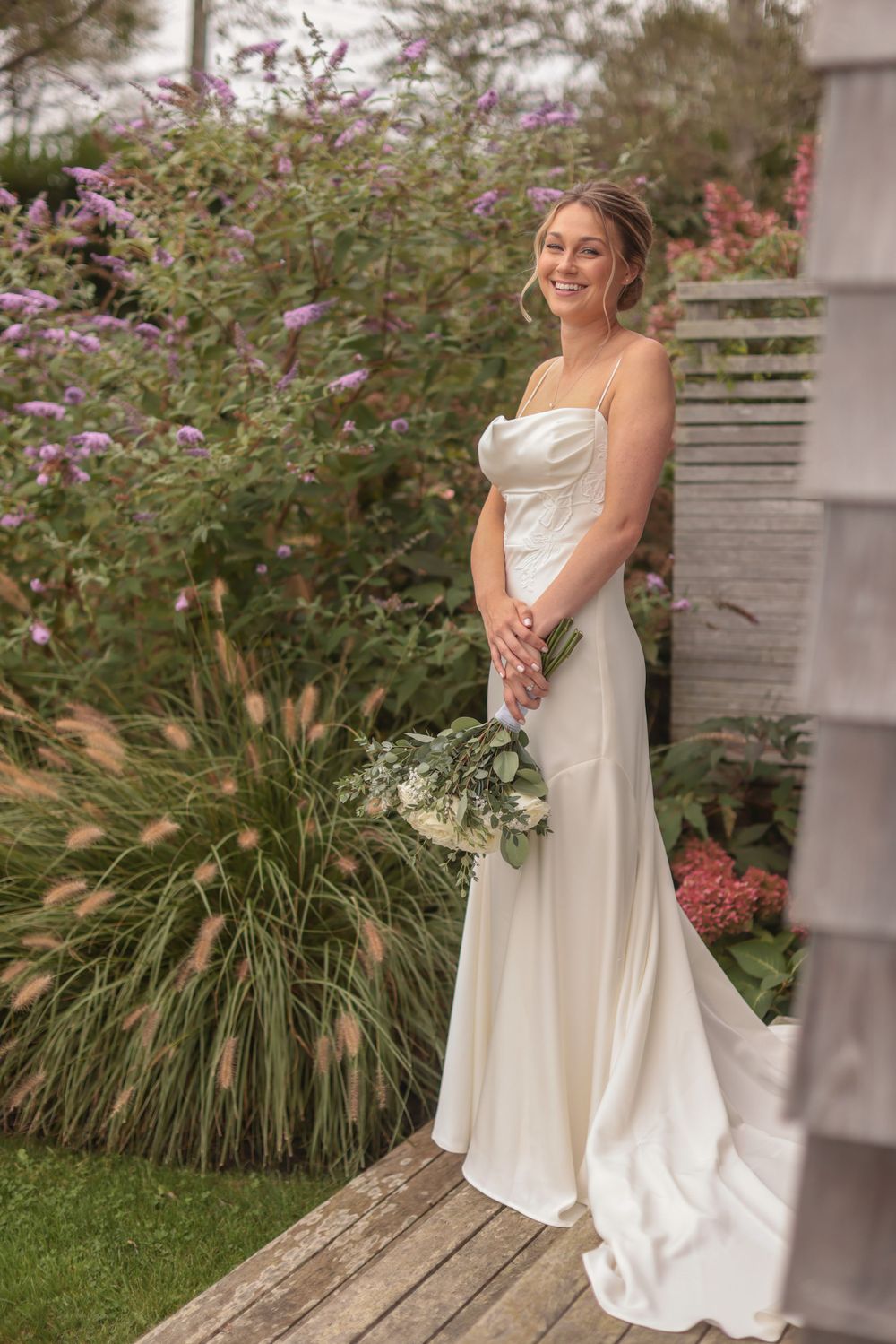 Bride in sleek white satin wedding dress standing on wooden deck surrounded by lush gardens.