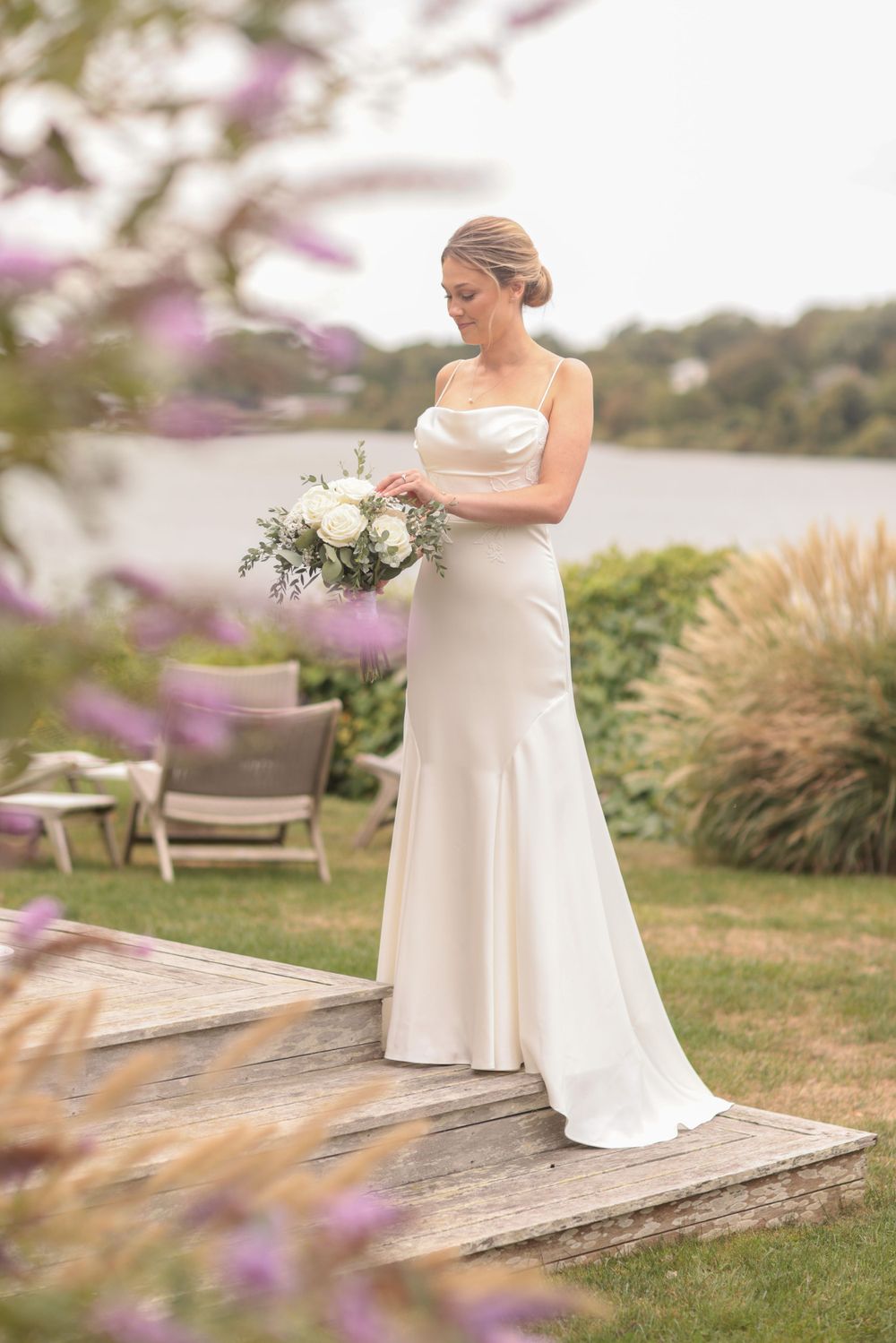 Elegant bride in white gown admiring her bouquet by a serene lakeside garden.