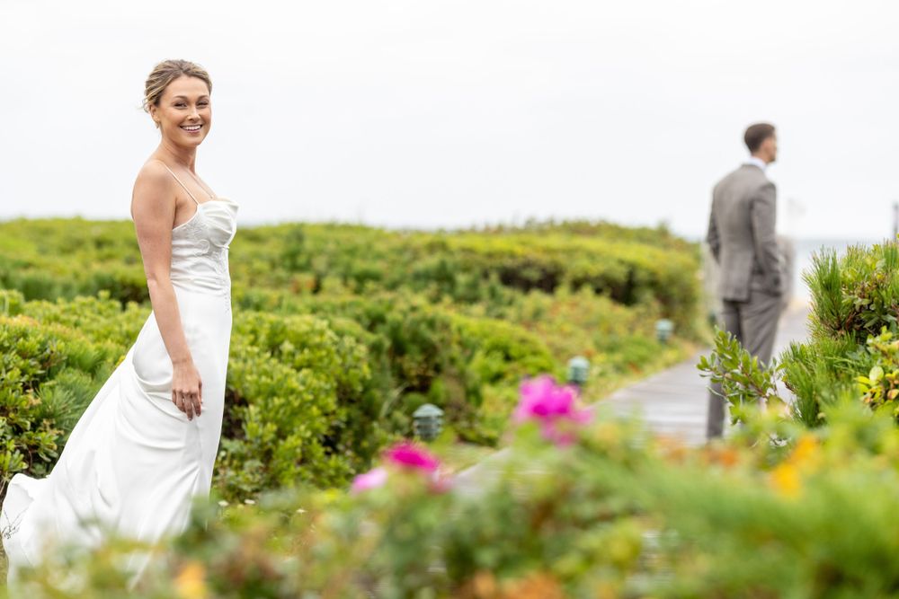 Bride in white gown smiling on a boardwalk path with groom in background amid greenery.