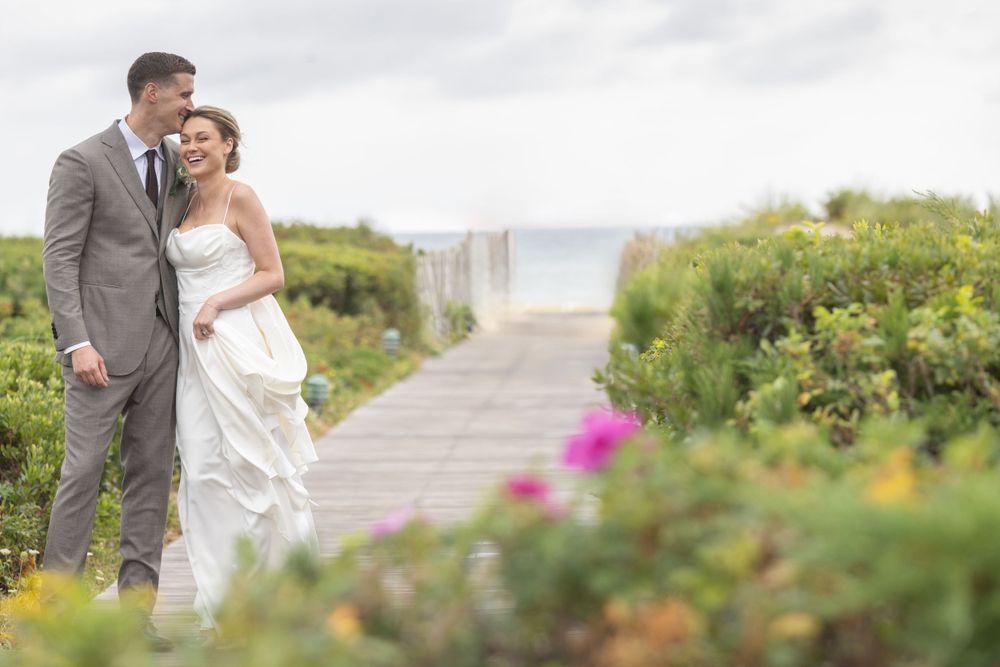Romantic couple embracing on a flower-lined boardwalk with ocean views in the background.