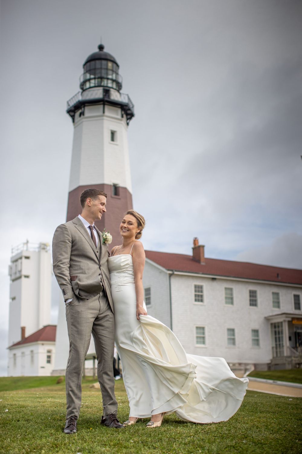 Bride and groom stand together on green grass in front of a tall historic lighthouse on a cloudy day.