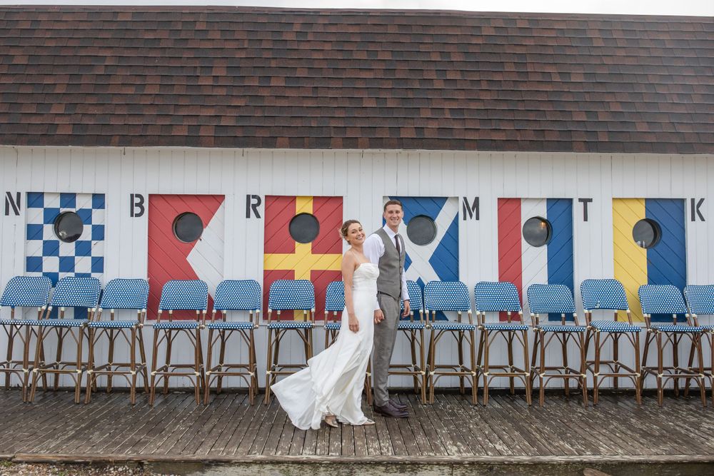 Bride and groom standing before a white boathouse wall decorated with colorful nautical signal flags.