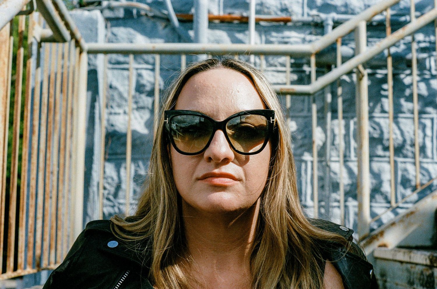 A person wearing sunglasses and a leather jacket stands in front of an industrial metal framework structure.