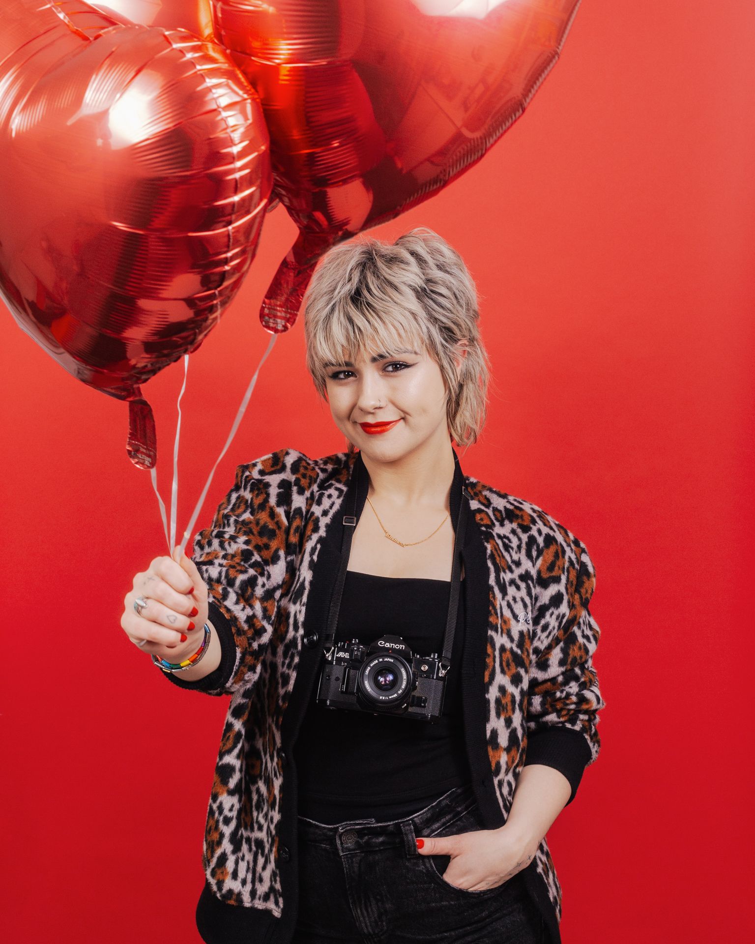A person wearing a leopard print jacket and holding red heart-shaped balloons poses against a vibrant red background.