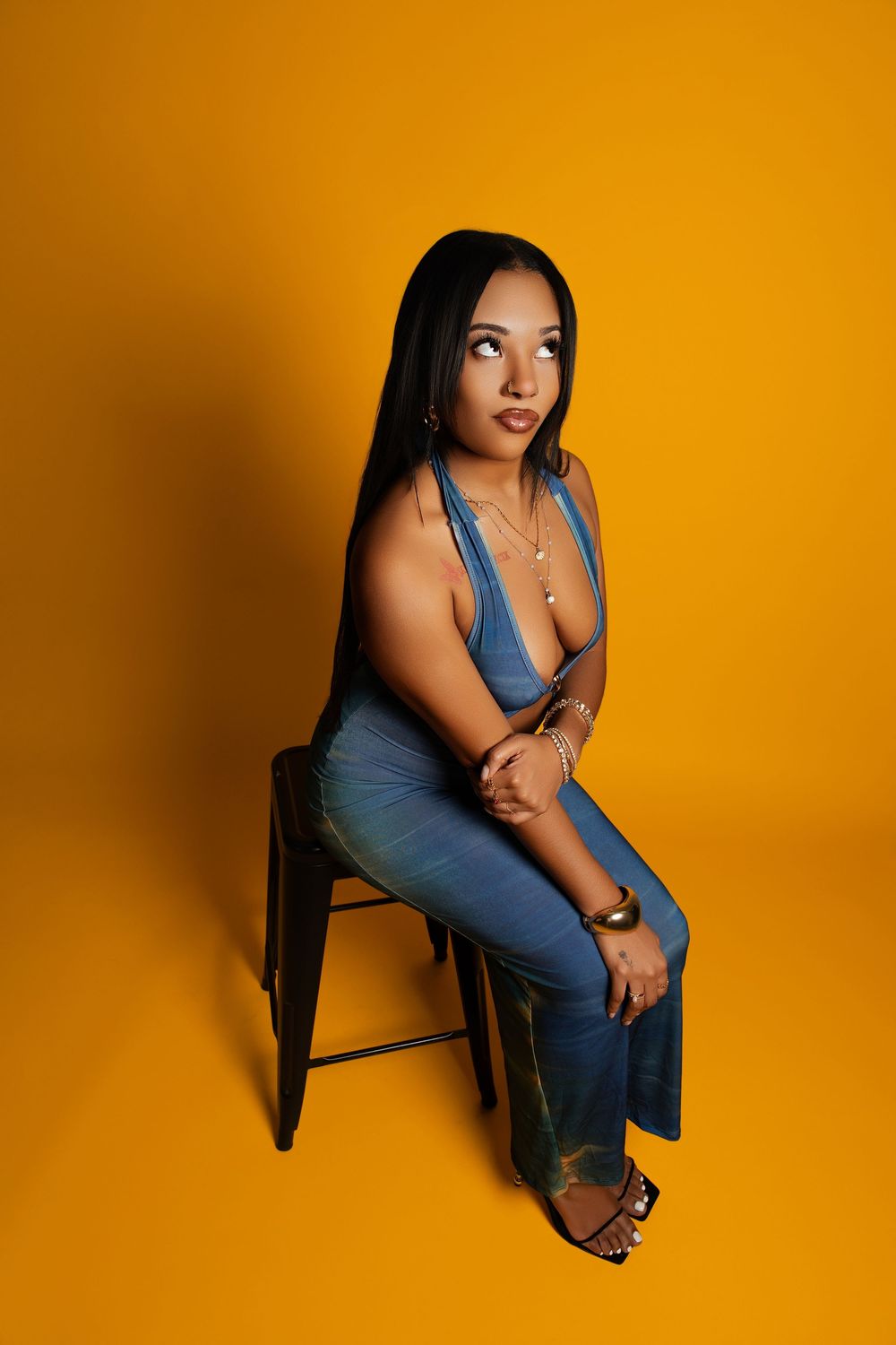 Model in blue dress poses artistically on chair against vibrant orange backdrop in studio photoshoot series.