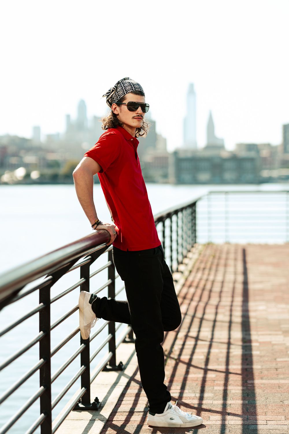 A person in a red shirt and black pants stretches and exercises on a waterfront railing with a city skyline in view.