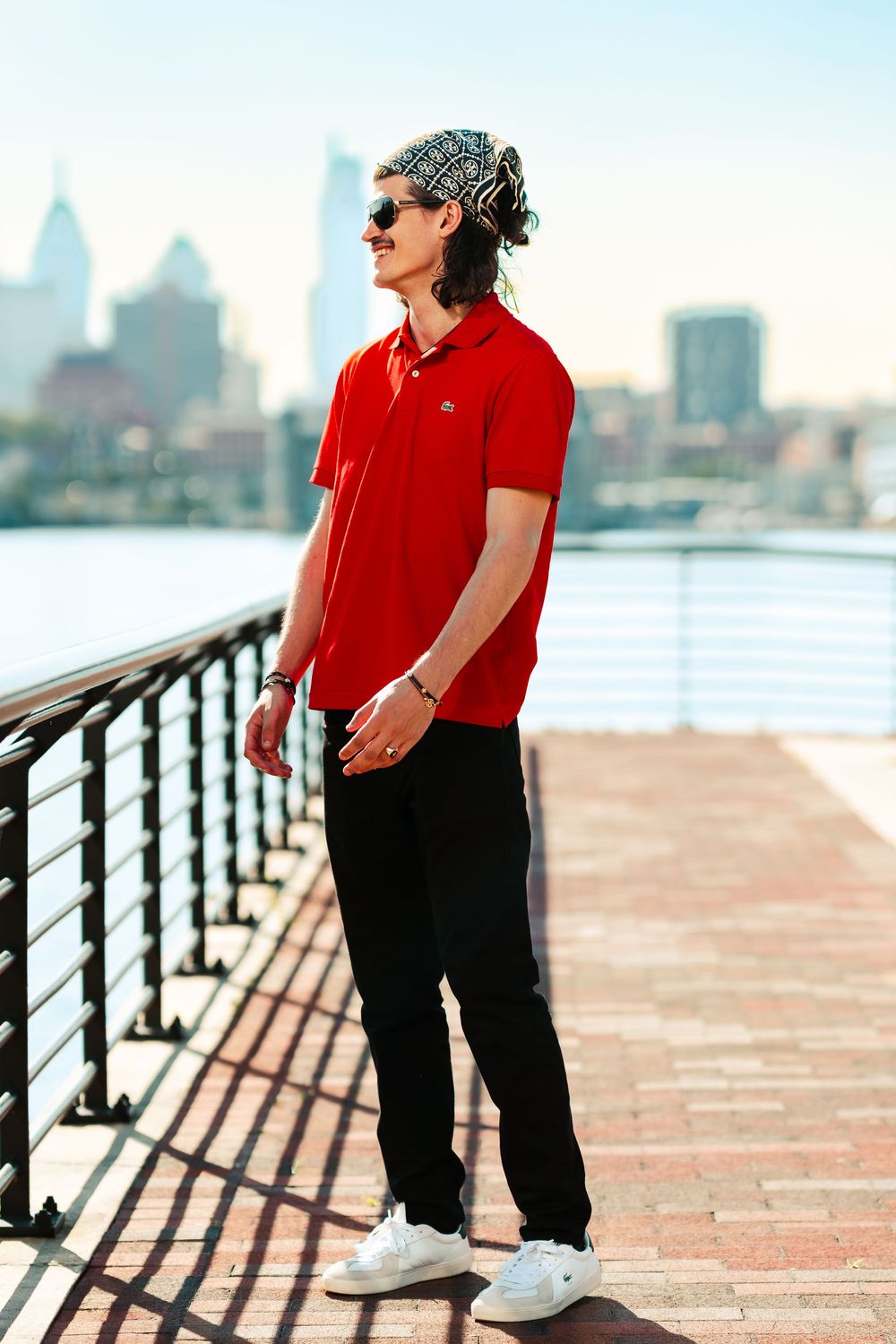 Person in red polo shirt and black pants poses on waterfront boardwalk with city skyline in background.