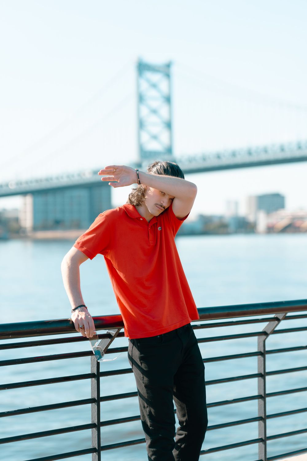 Person in red shirt leans against railing with Benjamin Franklin Bridge visible in background in Philadelphia.
