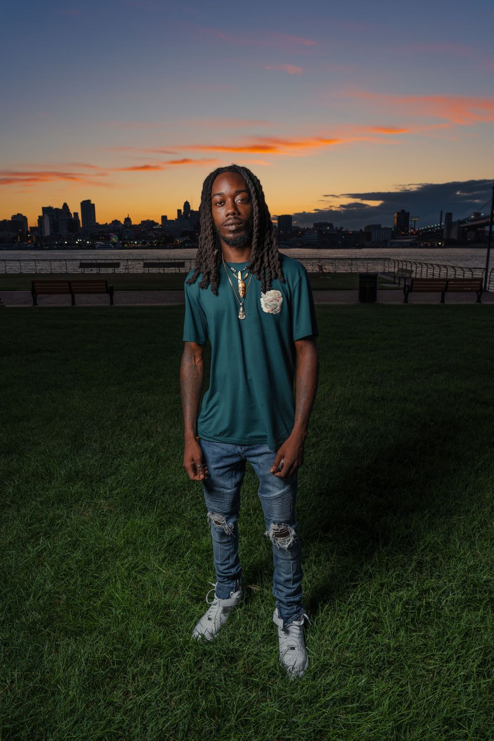 Person in teal shirt and ripped jeans stands in grassy field during sunset with city skyline in background.