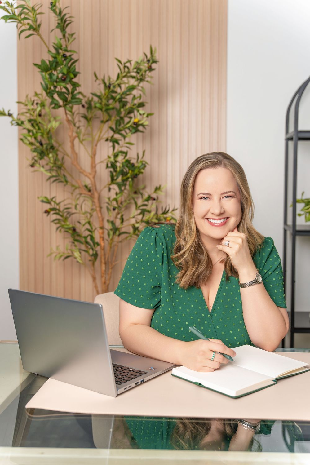 Professional at desk with laptop and notebook wearing green blouse in modern office with plants.