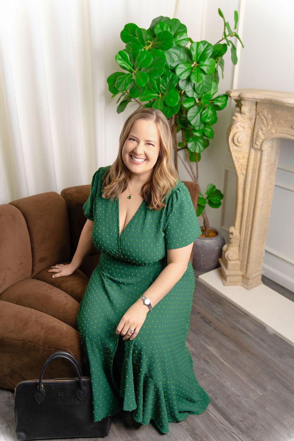 A woman in an emerald green polka dot dress poses on a brown couch in a bright interior space with houseplants.