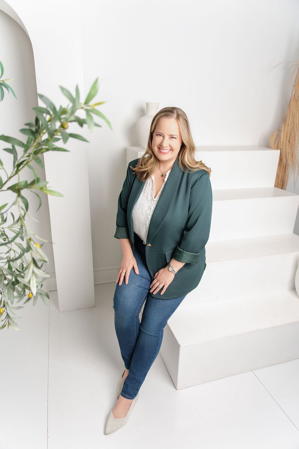Professional business portrait in white studio with green plants showing confident pose in blazer and jeans.