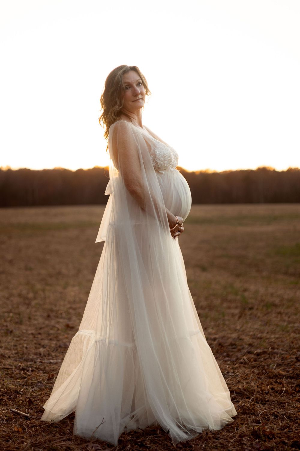 A pregnant woman stands ethereally in a field at sunset.