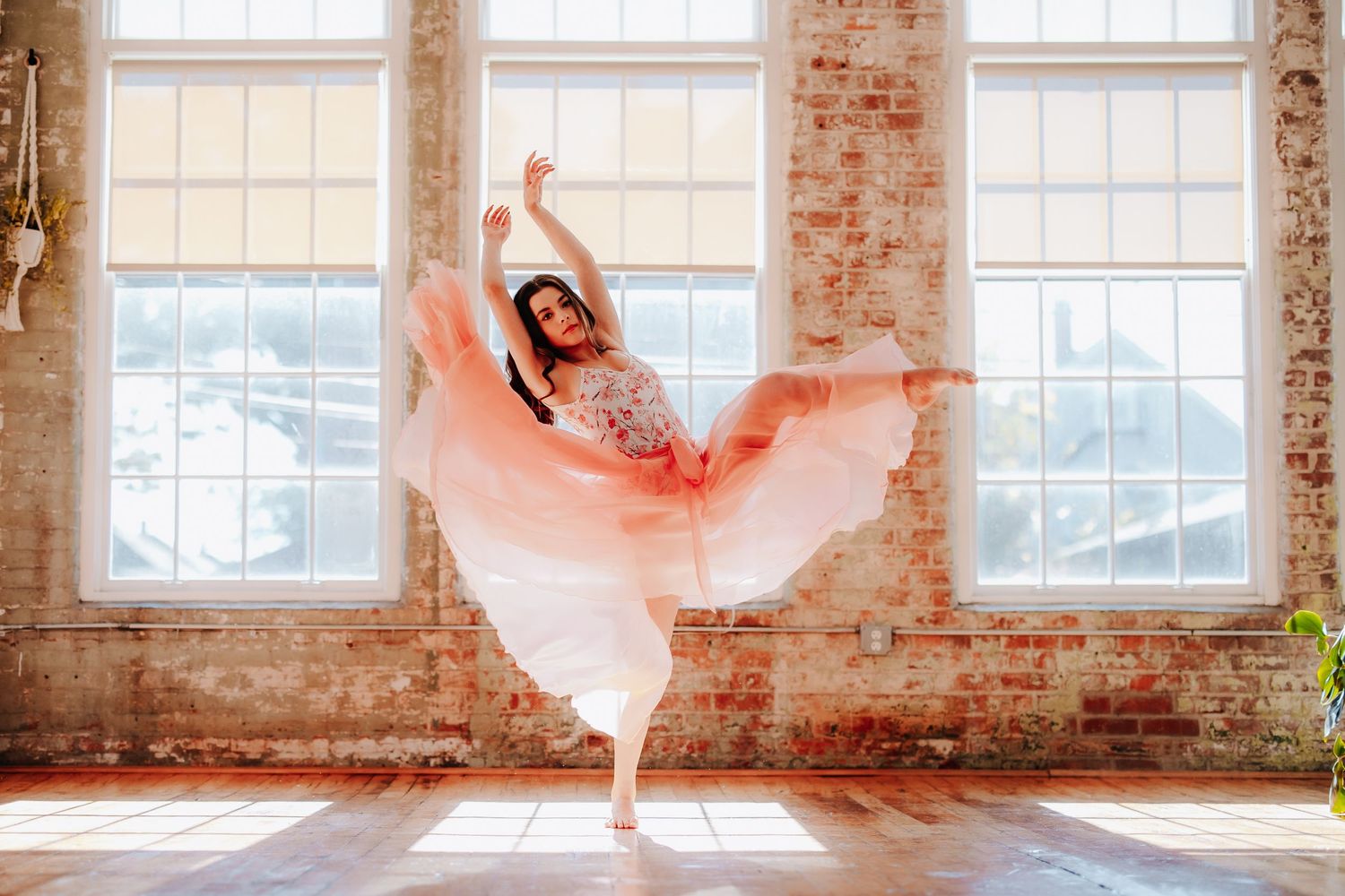 A dancer in a flowing pink dress leaps gracefully in front of sunlit industrial windows.