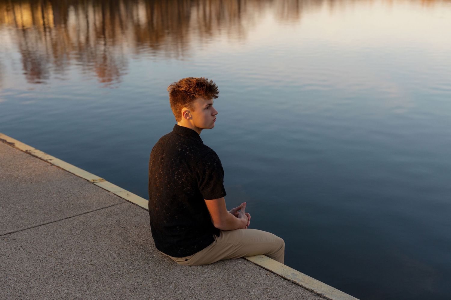 Someone in a black shirt sits peacefully at the edge of a calm lake during golden hour.