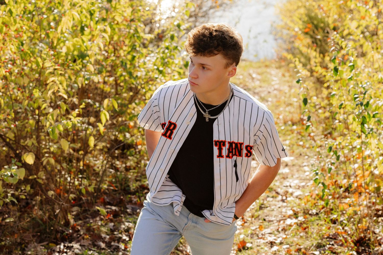 Baseball player in striped uniform poses outdoors against autumn foliage background in golden sunlight.