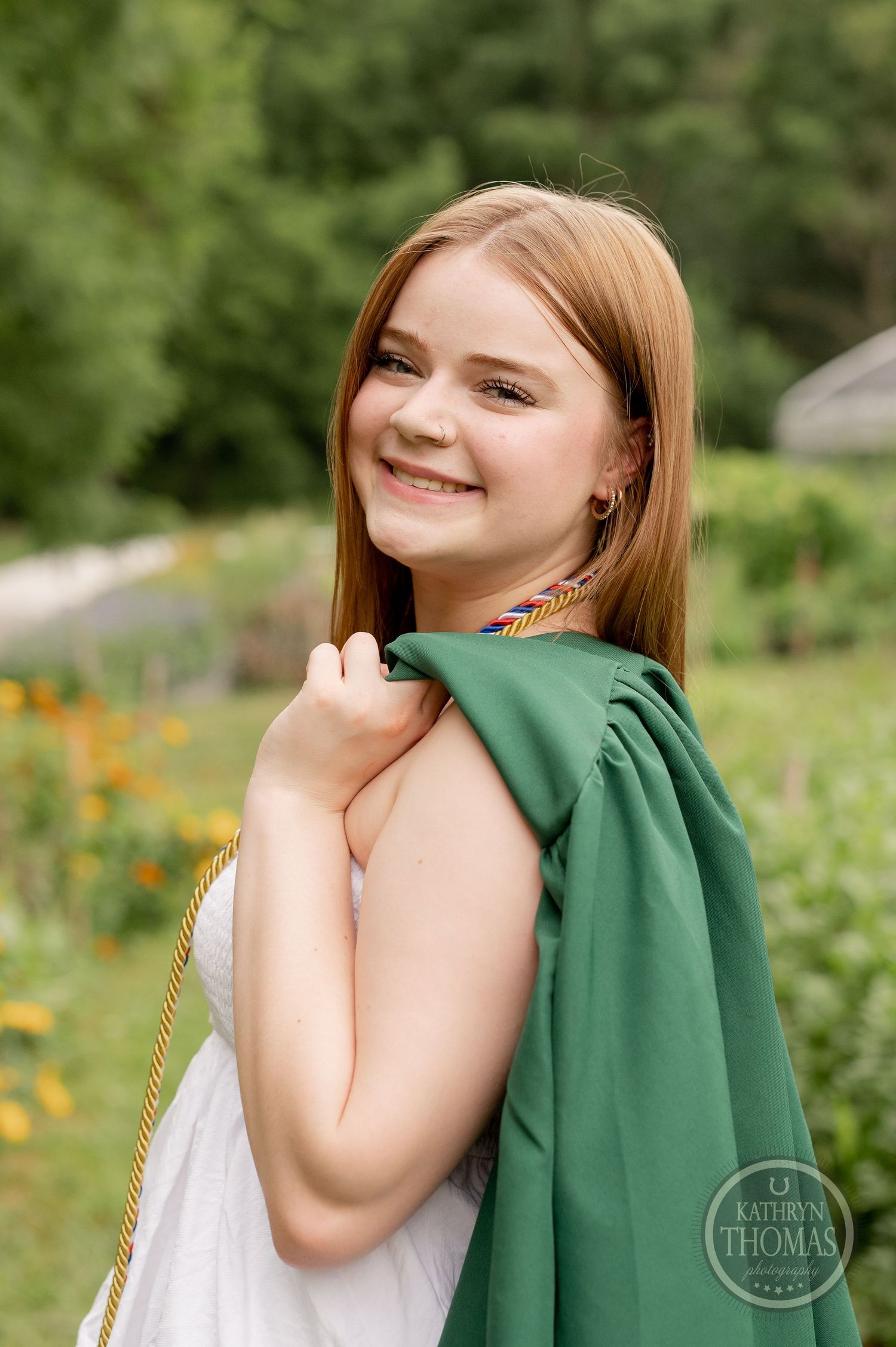 Graduate with green graduation gown draped over shoulder poses in garden setting.